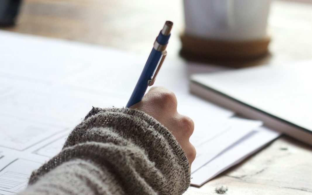 person writing on brown wooden table near white ceramic mug